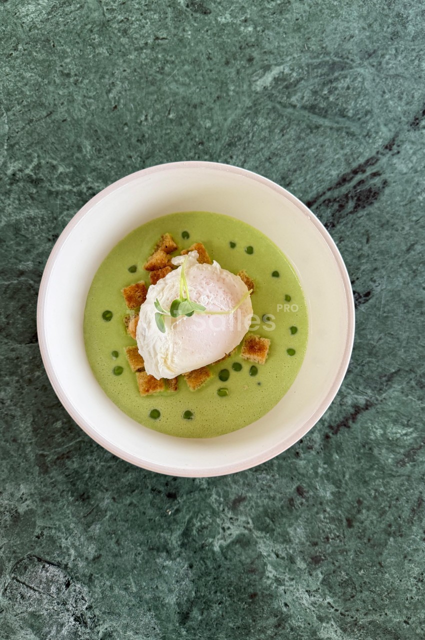 Bol de soupe verte garnie d'un œuf poché, de croûtons et d'une herbe fraîche, posé sur une table en marbre vert.