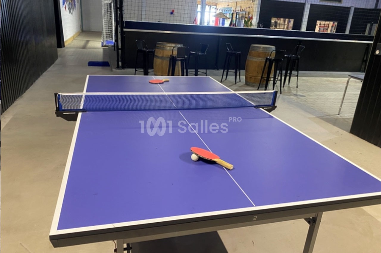Table de tennis de table bleue avec deux raquettes rouges posées, dans un espace intérieur avec des tables hautes en arrière…