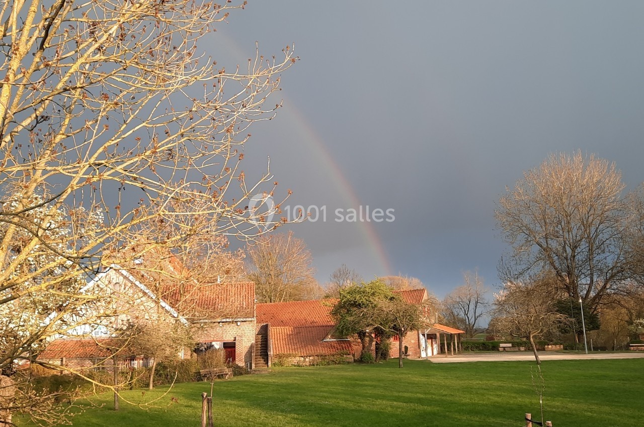 Un arc-en-ciel traverse un ciel nuageux au-dessus d'une maison en briques entourée d'arbres et d'une pelouse verte.