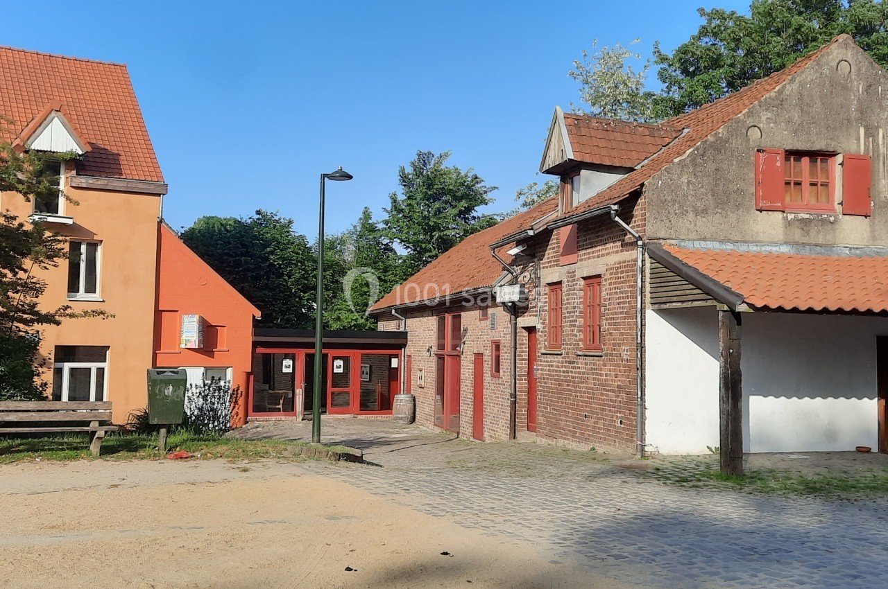 Bâtiments en briques rouges avec toits en tuiles, entourés d'arbres, sur une place pavée et ensoleillée.