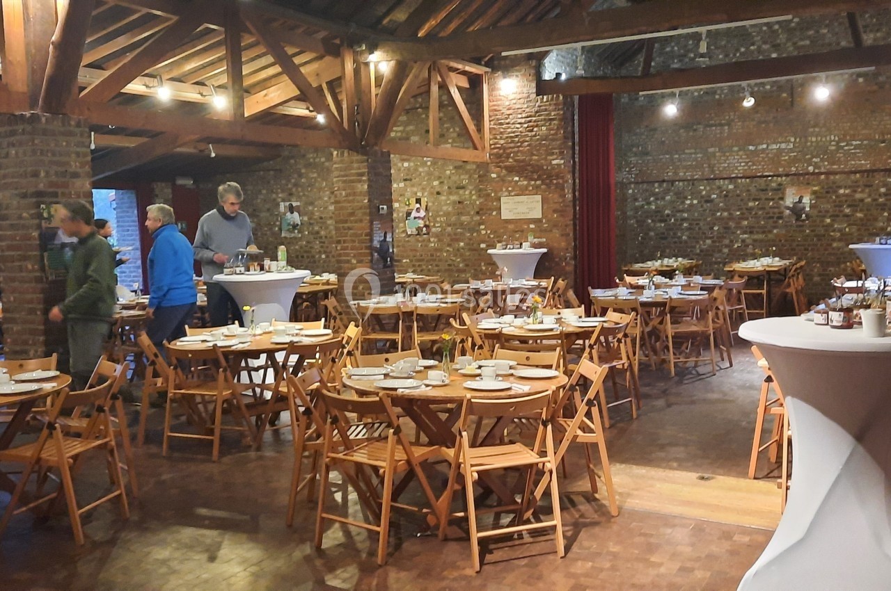 Salle en briques avec des tables rondes dressées pour un repas, quelques personnes en arrière-plan.