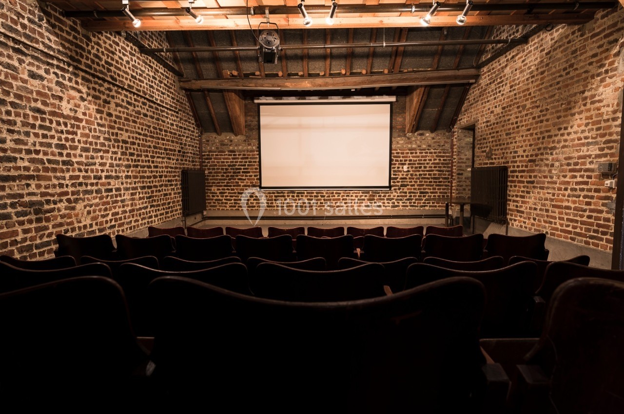 Salle de projection avec écran blanc, rangées de sièges en premier plan et murs en briques apparentes.