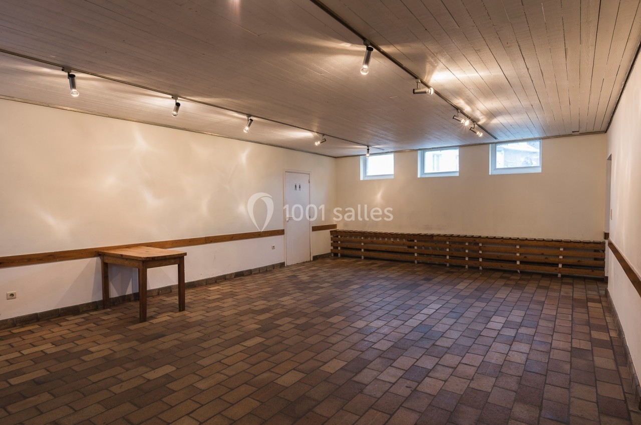 Salle vide avec murs blancs, plafond en bois, sol en carrelage et éclairage par spots, équipée d'une table et d'un banc.