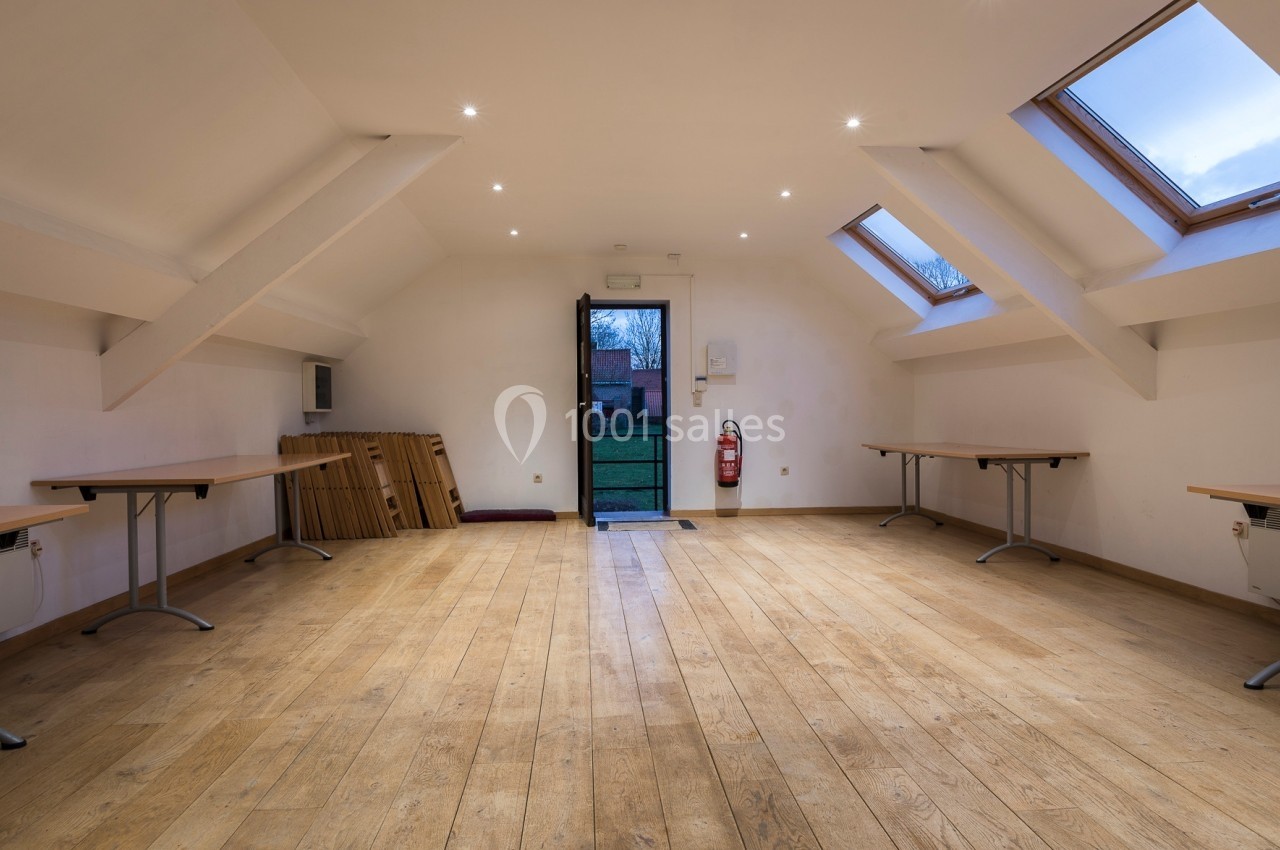Salle vide avec parquet en bois, tables pliées contre un mur, fenêtres de toit et porte ouverte donnant sur l'extérieur.