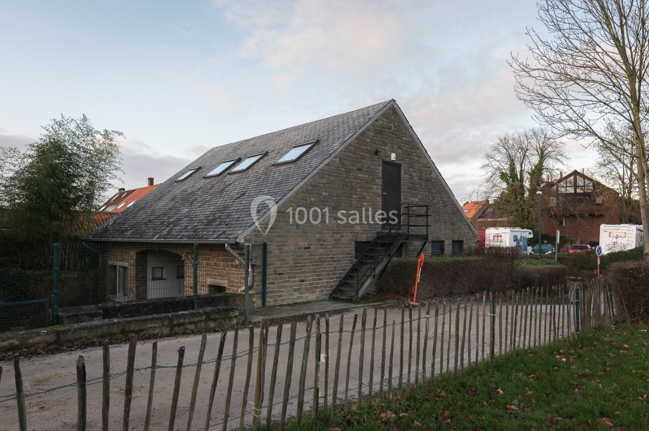 Bâtiment en pierre avec toit incliné, escaliers extérieurs et clôture en bois, entouré de végétation et de maisons.