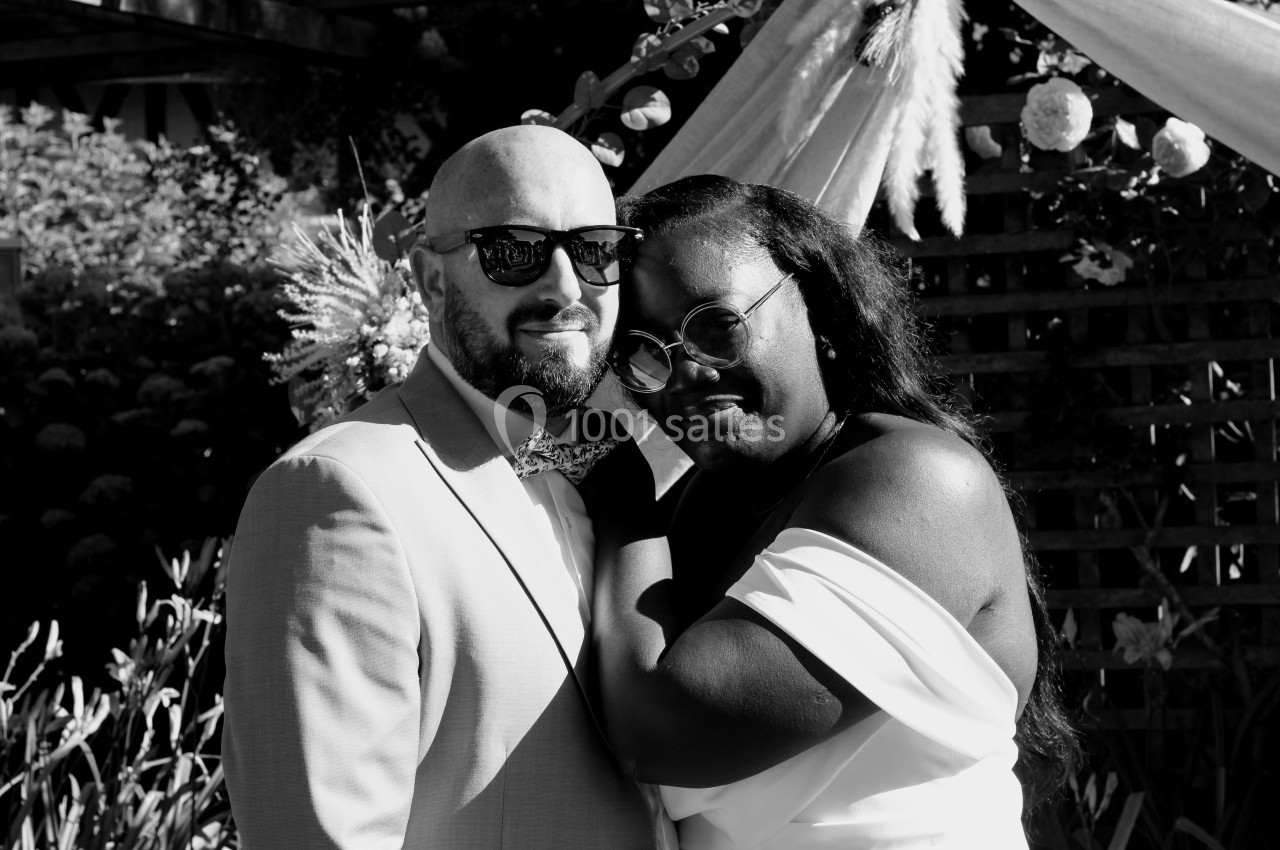 Un couple pose en noir et blanc devant une arche décorée de fleurs et de feuillages.