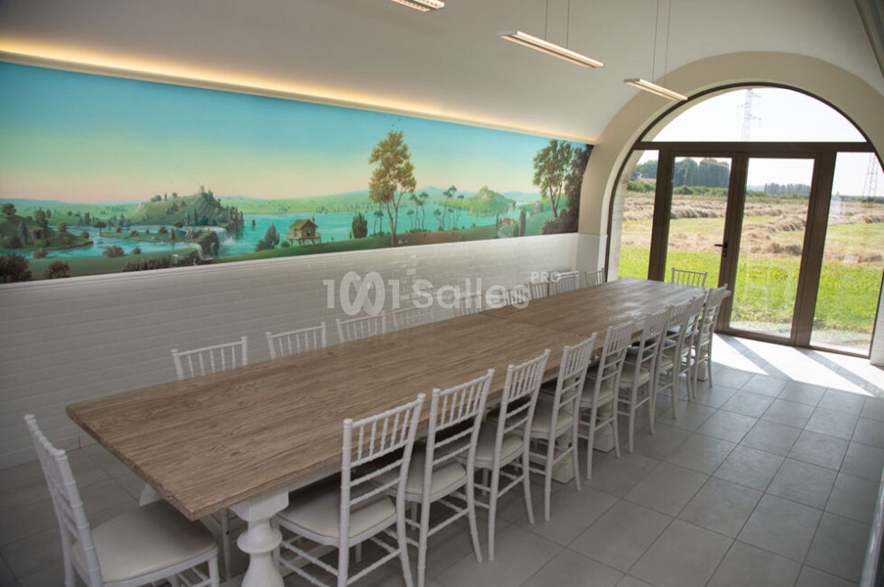 Grande table en bois entourée de chaises blanches dans une salle lumineuse avec fresque murale et vue sur un paysage…