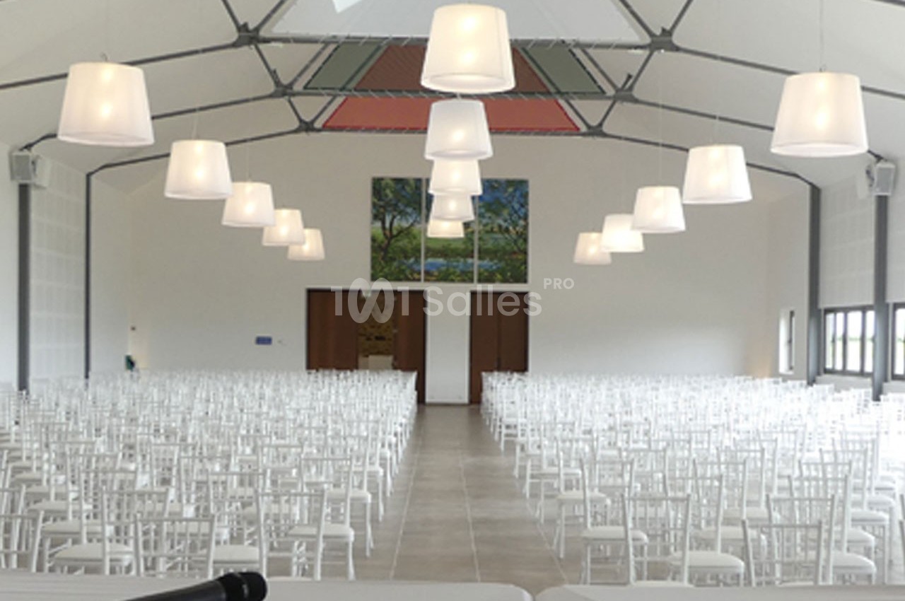 Salle de réception lumineuse avec des rangées de chaises blanches alignées sous des suspensions modernes.