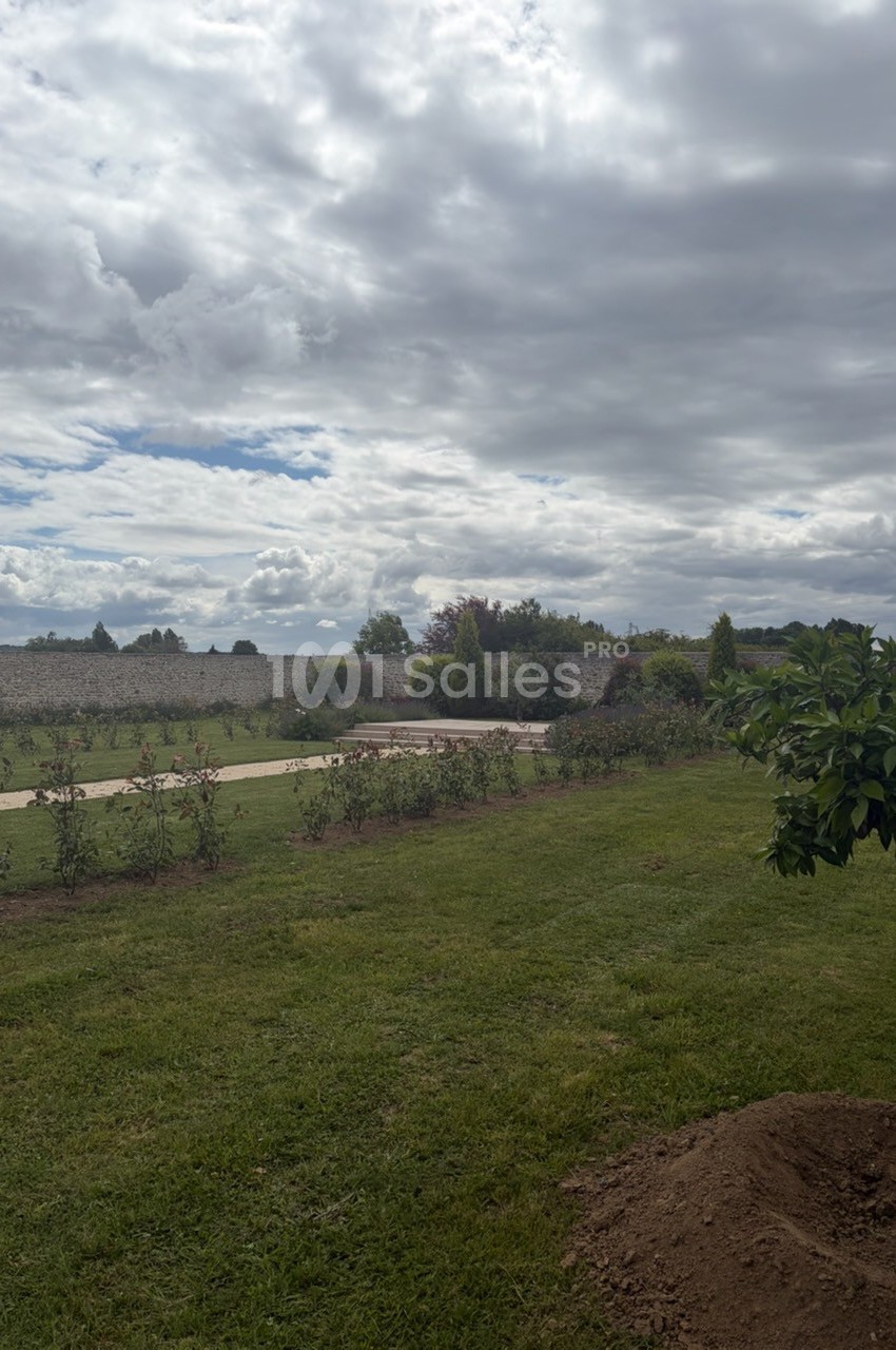 Jardin avec pelouse, allée bordée de rosiers, mur en pierre au fond et ciel nuageux.