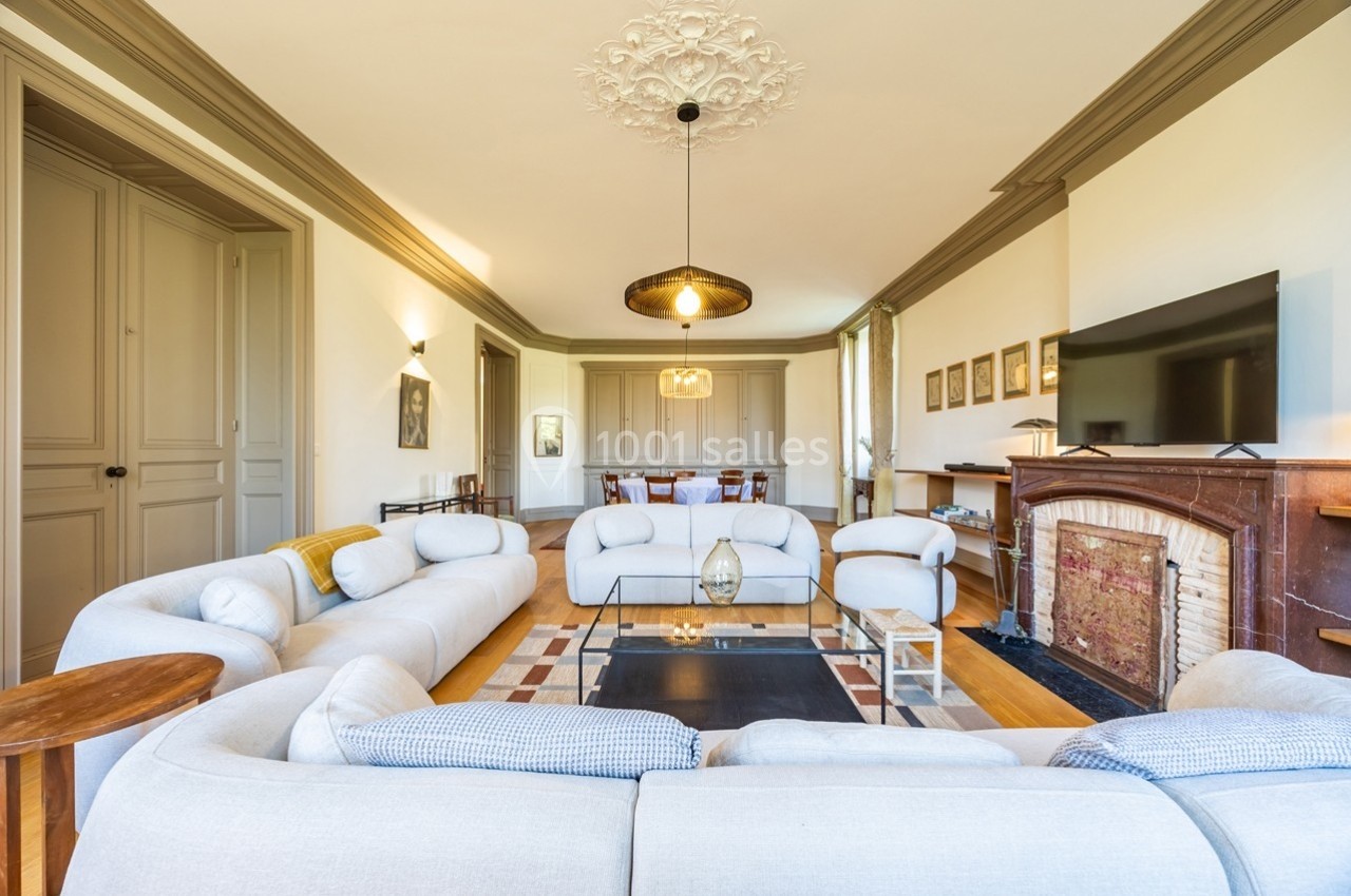 Salon lumineux avec canapés clairs disposés en U, table basse en verre, cheminée et télévision sur un mur beige.