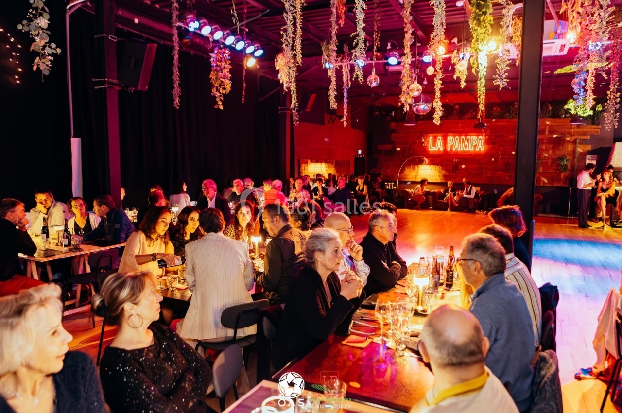 Salle animée avec des convives dînant à des tables éclairées, piste de danse et enseigne lumineuse ’La Pampa’ en arrière…