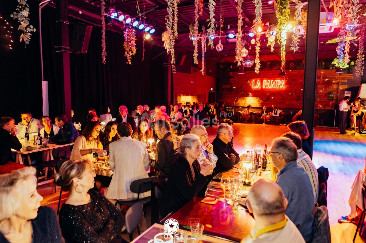 Salle animée avec des convives dînant à des tables éclairées, piste de danse et enseigne lumineuse ’La Pampa’ en arrière…