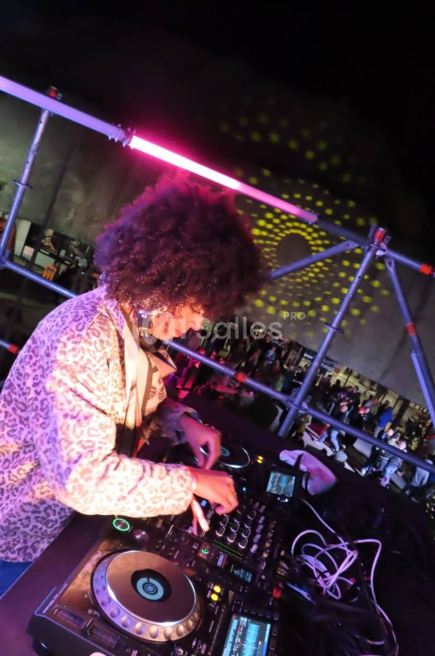 DJ mixant sur une scène en plein air, éclairée par des lumières colorées, avec un public en arrière-plan.