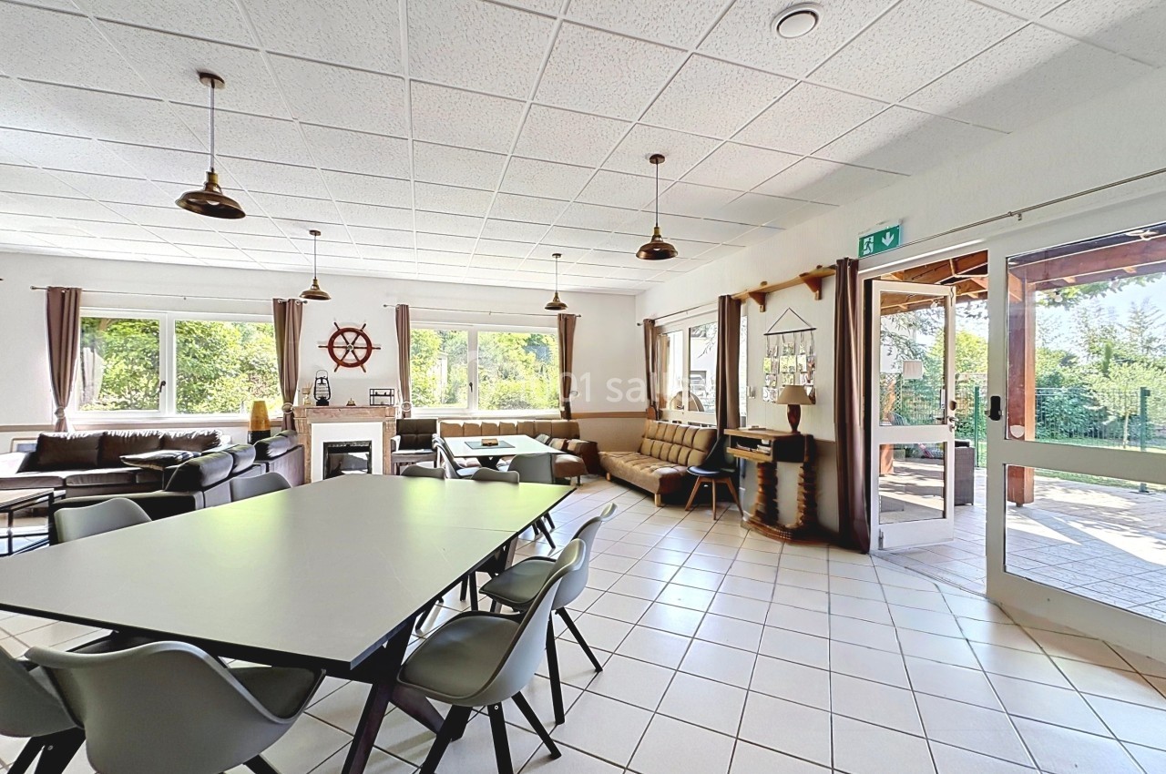 Salle lumineuse avec grandes fenêtres, tables, chaises, canapés et décorations nautiques, donnant sur un espace extérieur.