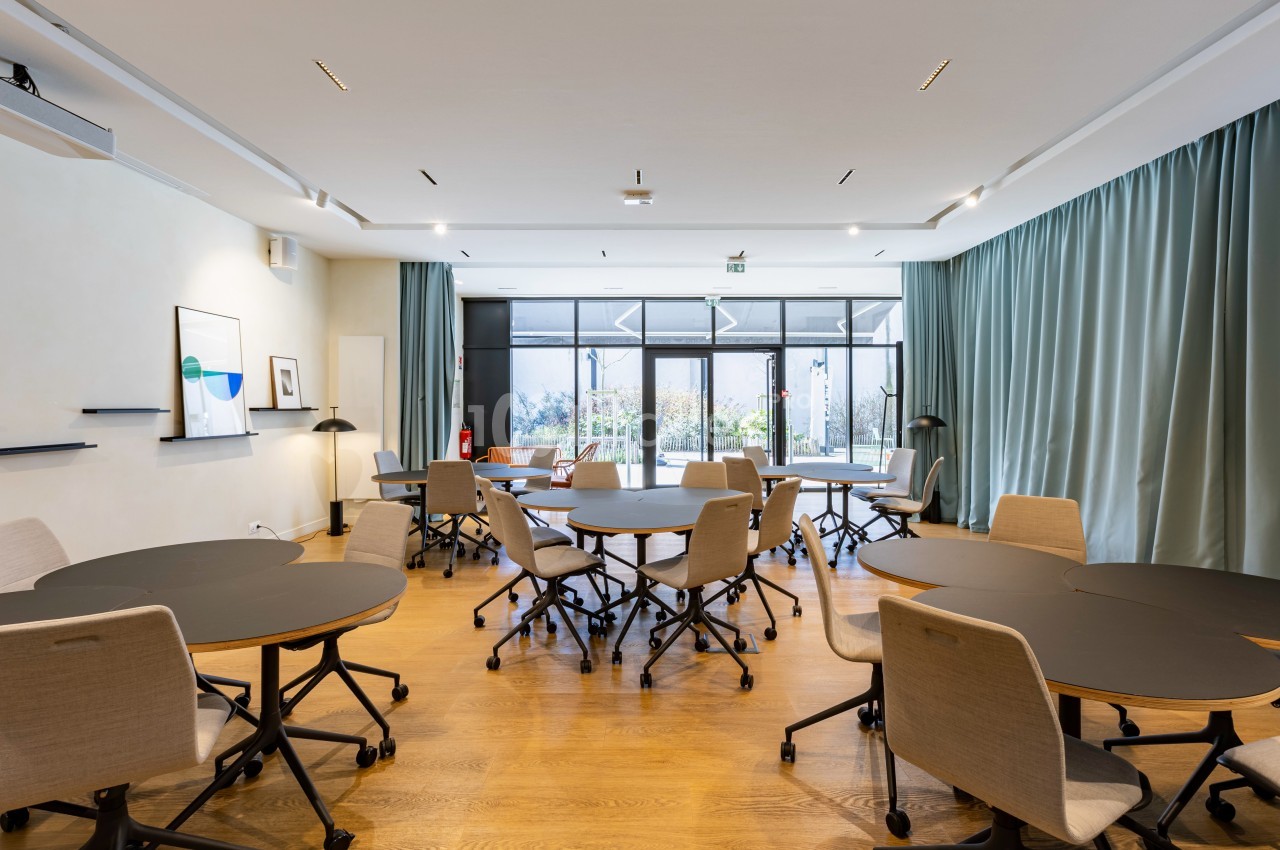 Salle lumineuse avec plusieurs tables rondes noires entourées de chaises, grandes baies vitrées et rideaux verts.