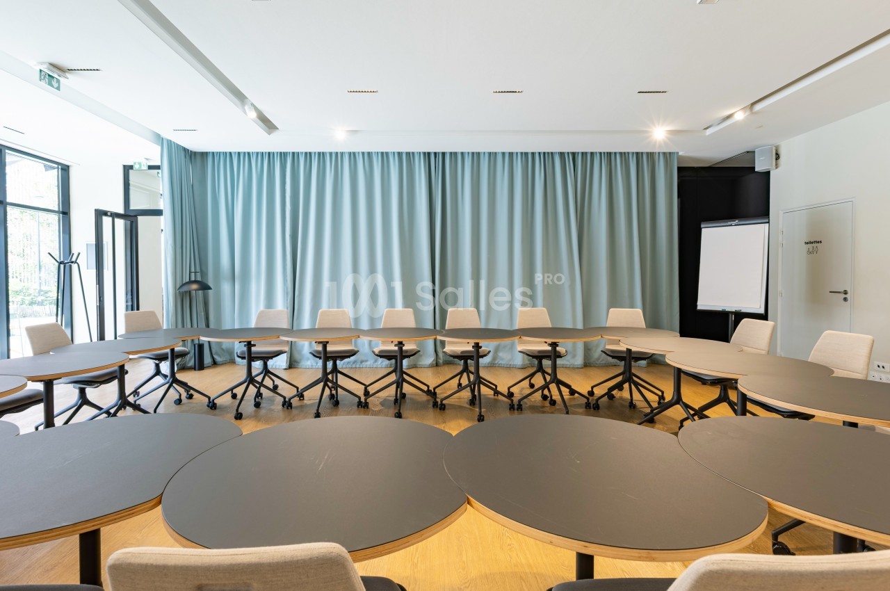 Salle de réunion lumineuse avec tables ovales noires, chaises à roulettes et rideaux bleus en arrière-plan.