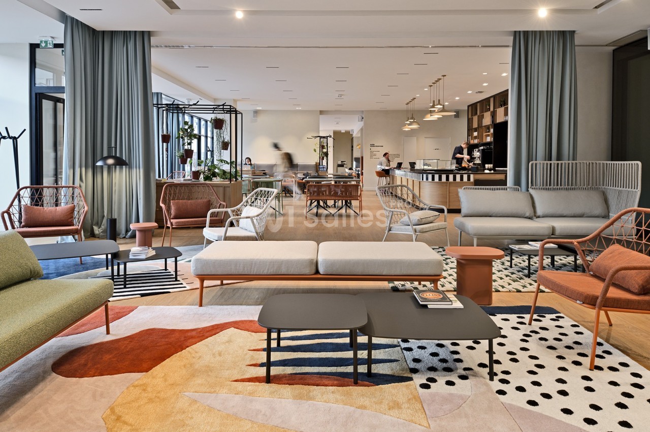 Espace lounge moderne avec canapés, fauteuils et tables basses, décoré de tapis colorés et plantes en arrière-plan.