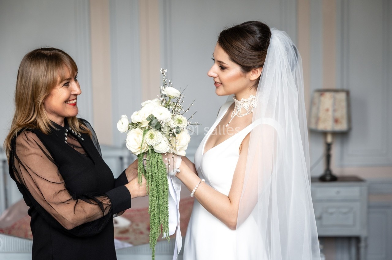 Une mariée en robe blanche et voile discute avec une femme tenant un bouquet de fleurs dans une pièce élégante.