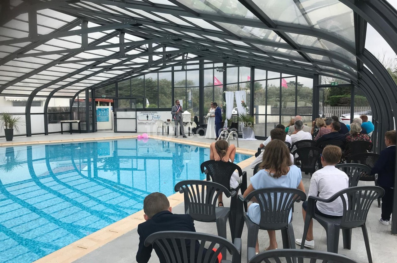 Cérémonie près d'une piscine couverte, avec des invités assis et un couple debout devant une table décorée.