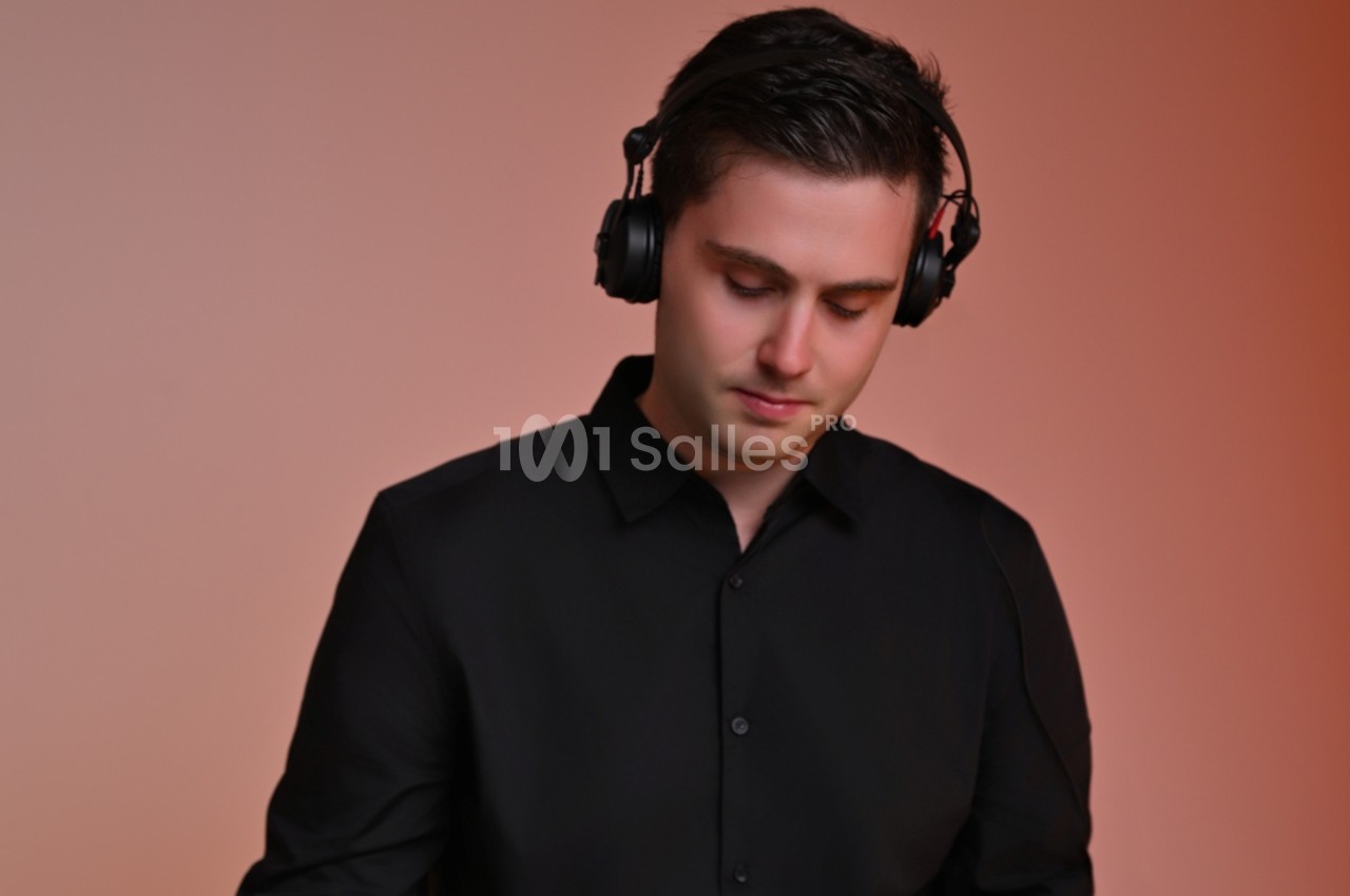 Un homme portant un casque audio noir, vêtu d'une chemise noire, se tient devant un fond dégradé orange.