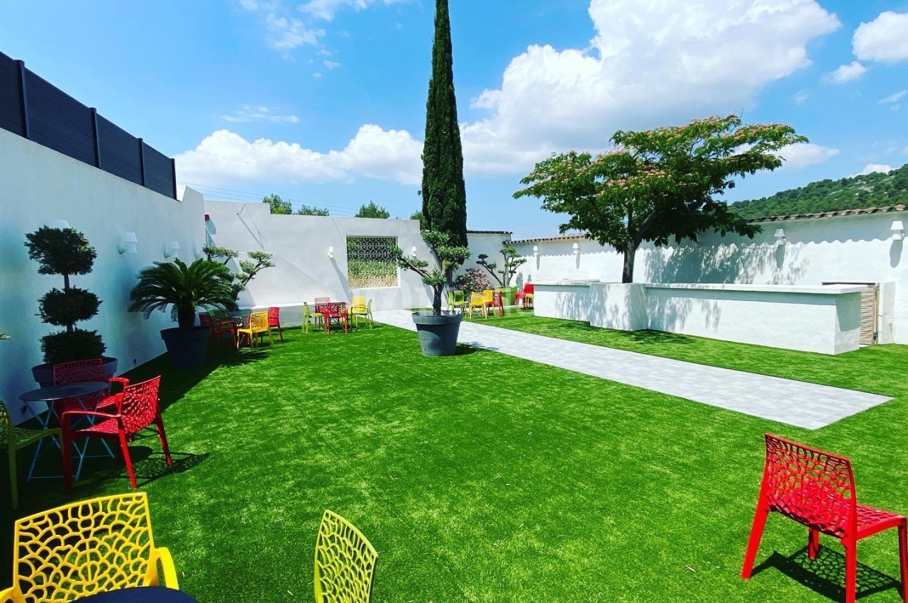 Jardin avec pelouse, chaises colorées, tables noires, un cyprès central et un ciel bleu en arrière-plan.
