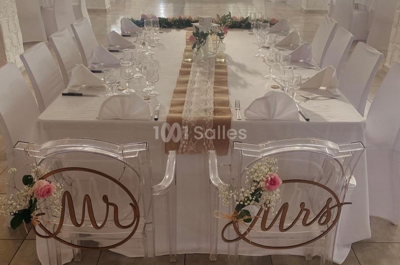 Table de mariage décorée avec nappes blanches, chemins de table en dentelle et chaises transparentes ornées des mots ’Mr’ et…