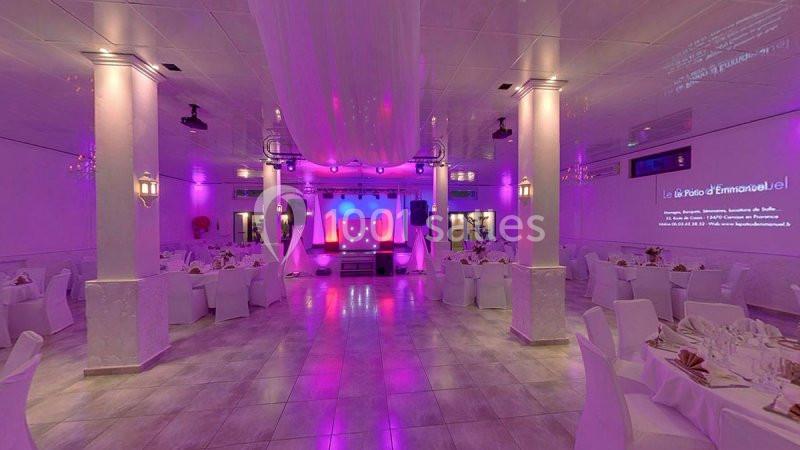 Salle de réception décorée avec des tables dressées, éclairage violet et scène équipée de matériel audiovisuel.