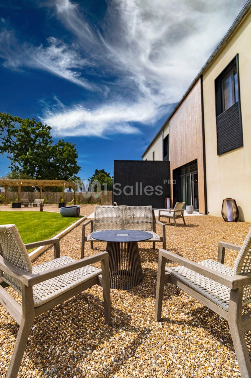 Terrasse avec mobilier de jardin en bois et métal sur gravier, entourée de pelouse et d'un bâtiment moderne.