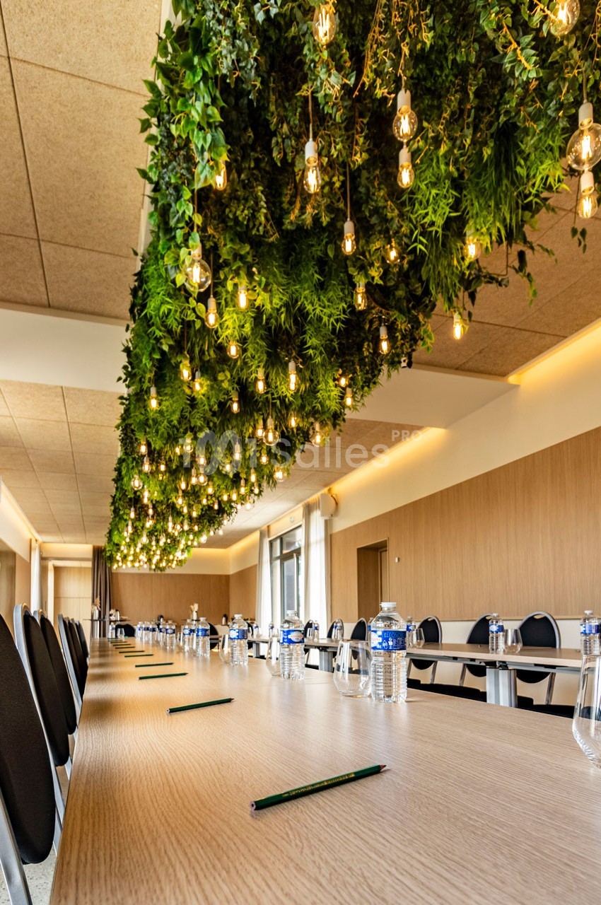 Salle de réunion avec une longue table en bois, des chaises noires et un plafond décoré de plantes et de lumières suspendues.