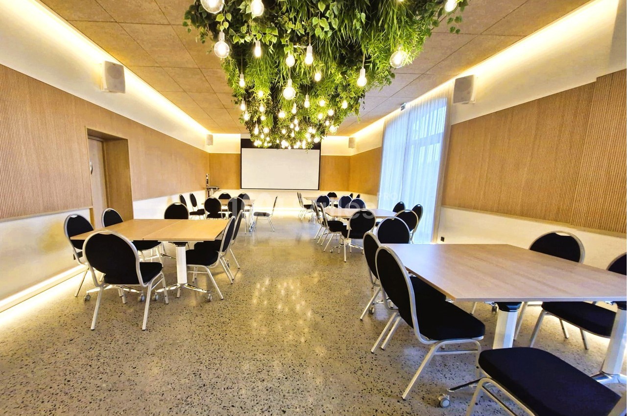 Salle de réunion lumineuse avec tables rectangulaires, chaises noires et plafond décoré de plantes et ampoules suspendues.