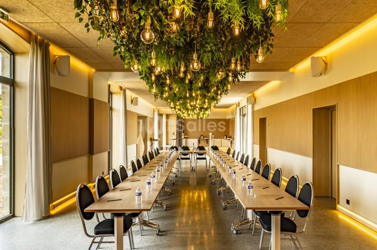 Salle de réunion lumineuse avec une longue table en U, des chaises noires et un plafond décoré de plantes suspendues.