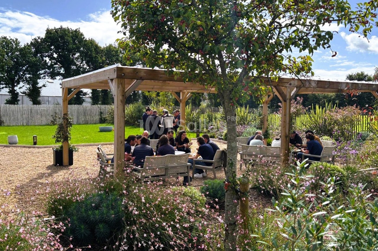 Un groupe de personnes assises sous une pergola en bois dans un jardin fleuri par une journée ensoleillée.