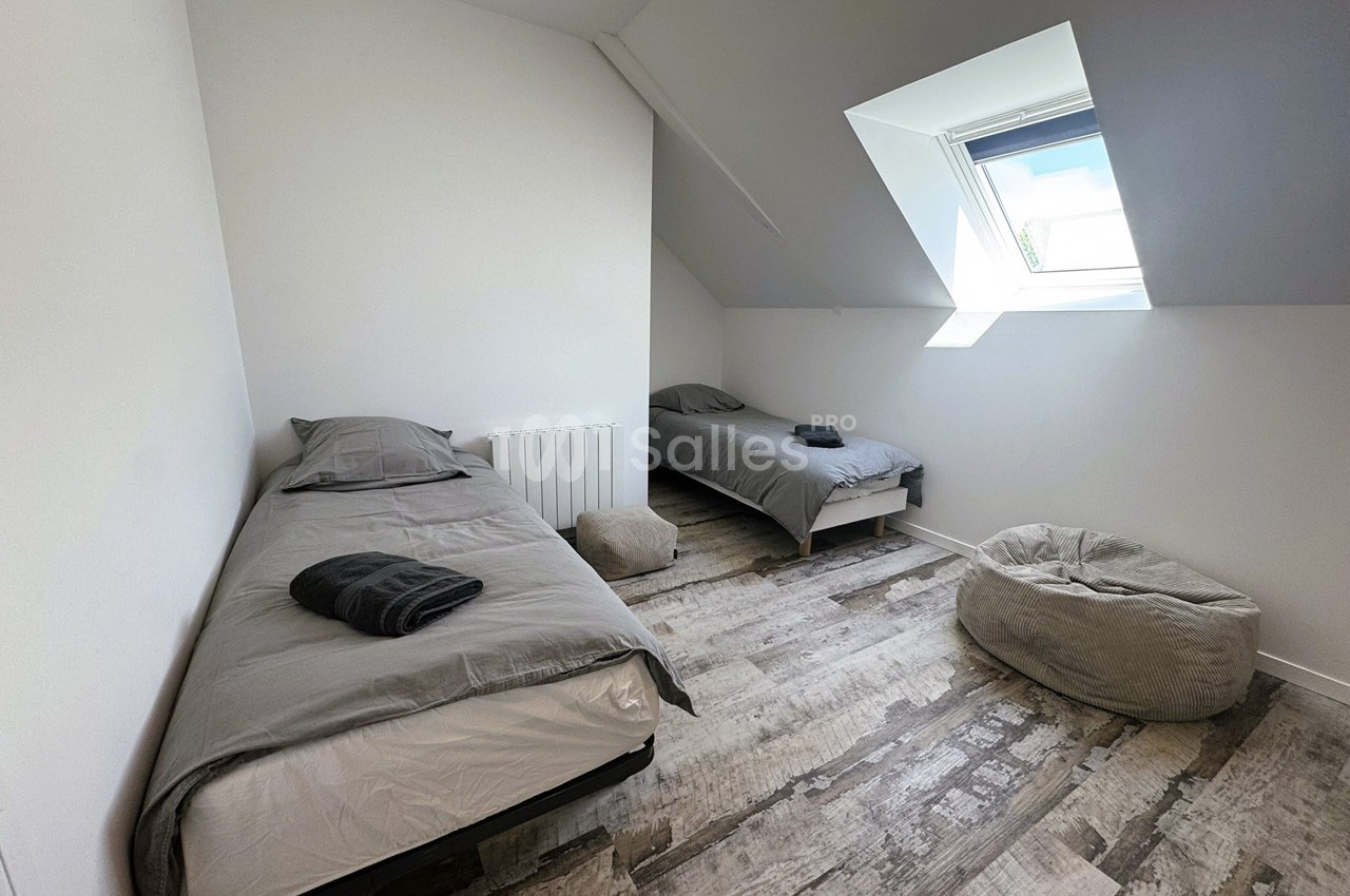 Deux lits simples avec draps gris dans une chambre lumineuse sous les combles, avec un pouf et un sol en bois clair.