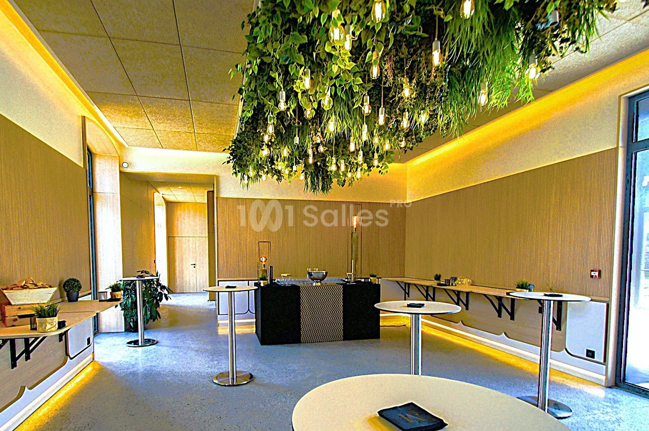 Salle lumineuse avec tables hautes, décor végétal suspendu et éclairage chaleureux.