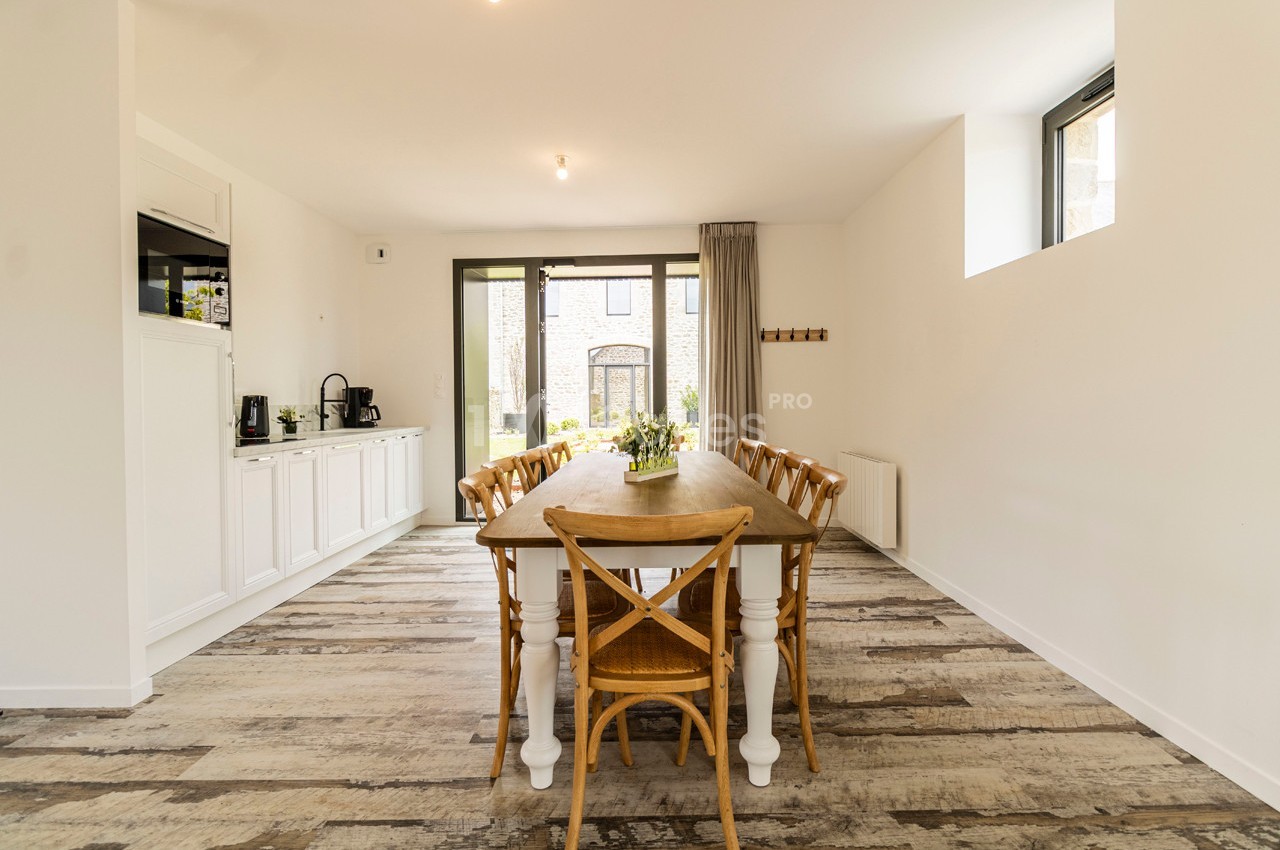 Salle à manger lumineuse avec une grande table en bois entourée de chaises, sol en parquet et mobilier blanc.