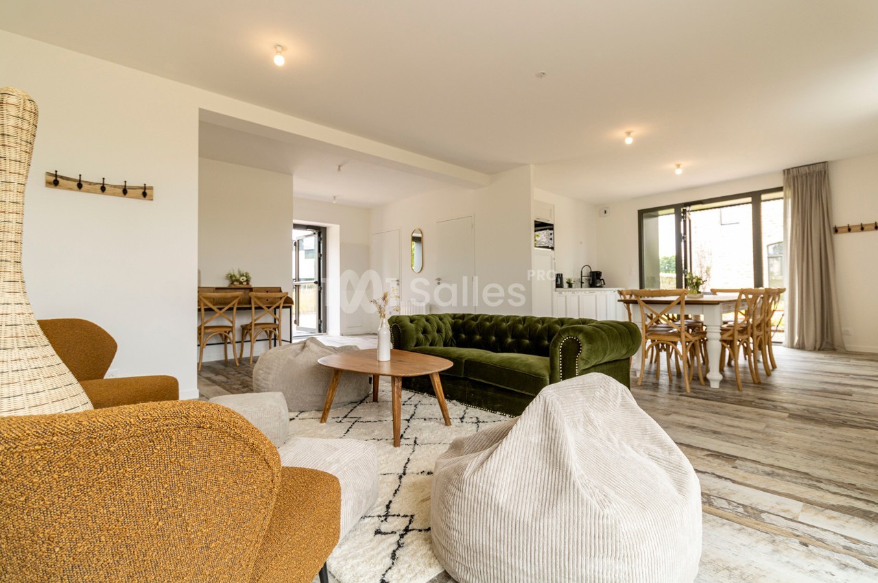 Salon spacieux avec canapé vert, fauteuils, poufs, table basse et coin repas, éclairé par de grandes fenêtres.