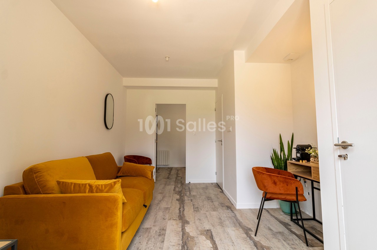 Salon lumineux avec canapé jaune, bureau en bois, chaise orange, plante verte et sol en parquet clair.