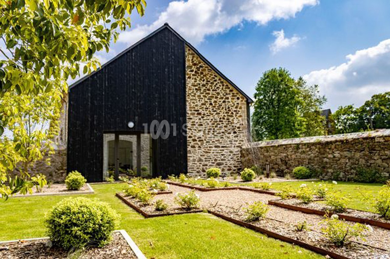 Façade d'une maison en pierre et bois noir, entourée d'un jardin aménagé avec des parterres et des allées gravillonnées.