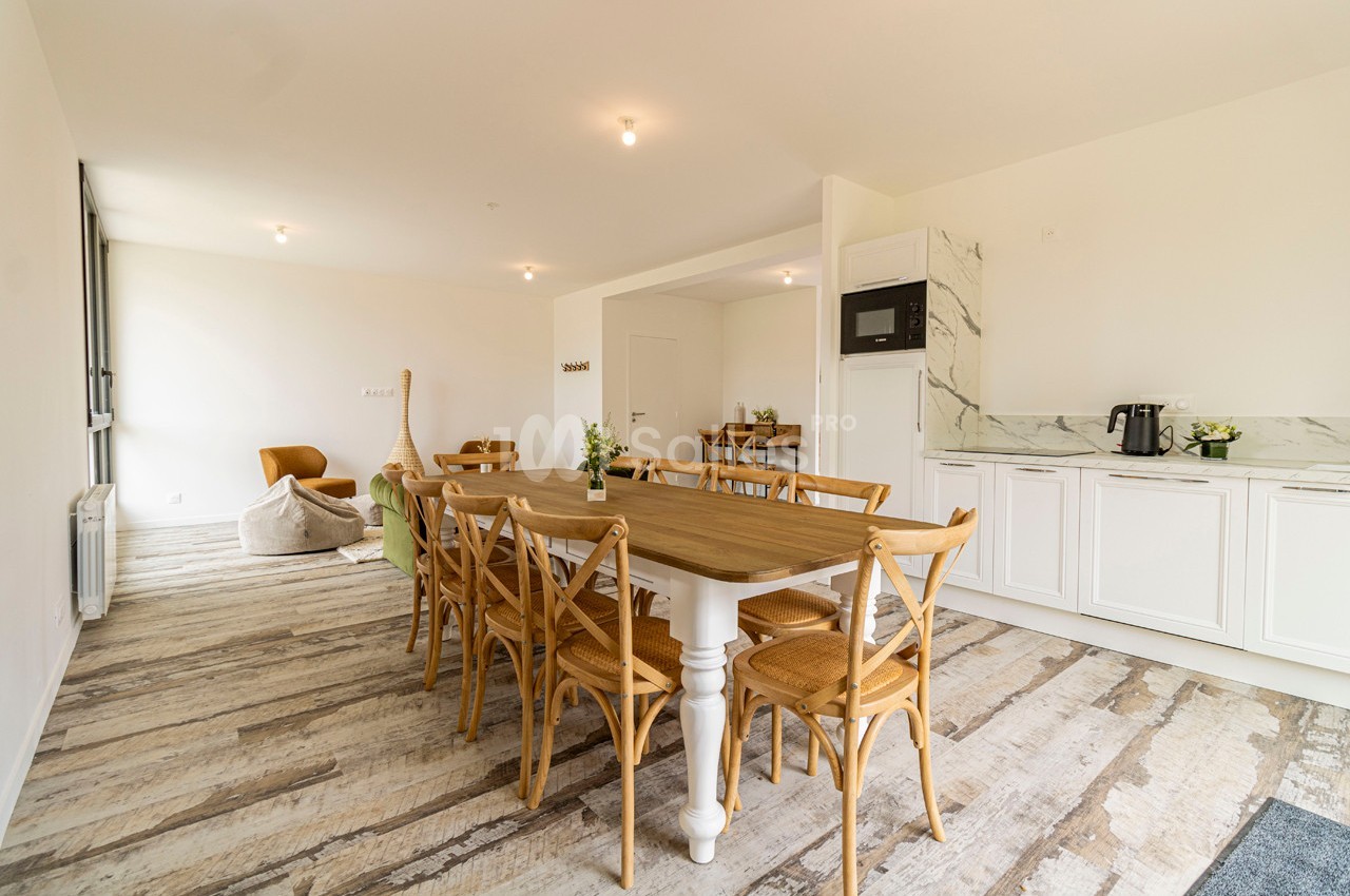 Salle à manger lumineuse avec une grande table en bois, chaises en bois clair et cuisine ouverte équipée en arrière-plan.