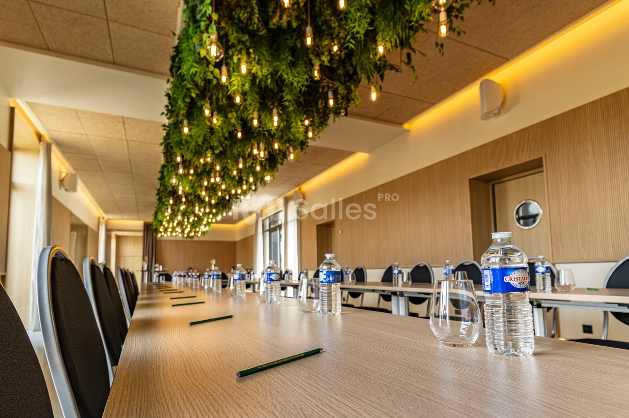 Salle de réunion avec une longue table en bois, des bouteilles d'eau, des verres, des stylos et un plafond décoré de plantes…