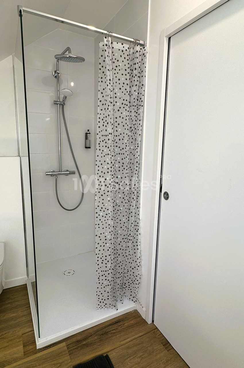 Douche moderne avec paroi vitrée, rideau à motifs noirs, sol en bois et porte blanche adjacente.