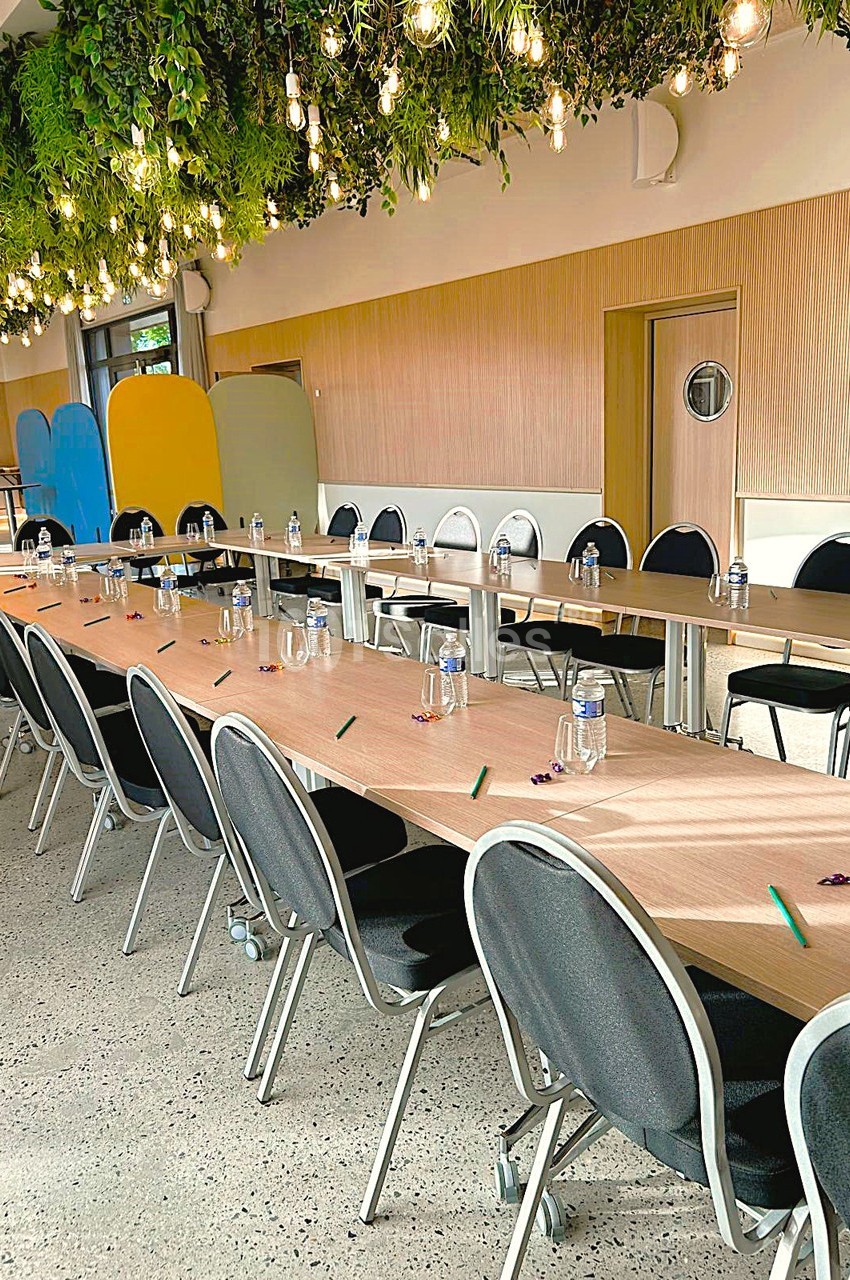 Salle de réunion avec longues tables alignées, chaises noires, bouteilles d'eau et crayons, sous un plafond végétalisé.