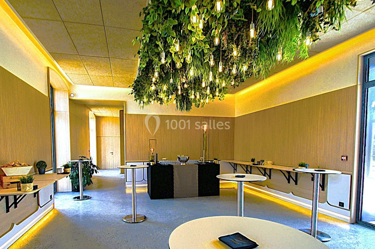 Salle lumineuse avec tables hautes, plafond décoré de plantes suspendues et éclairage chaleureux.