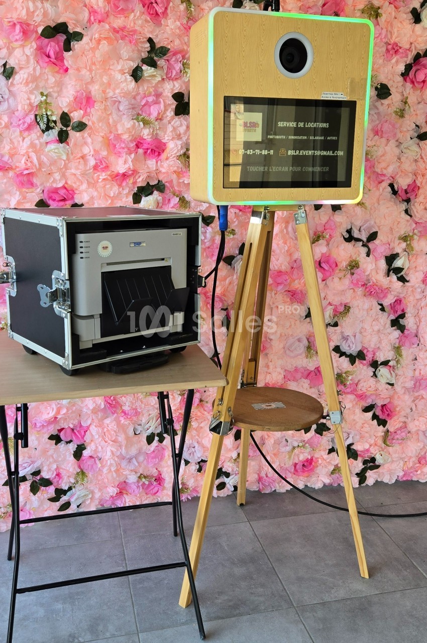 Borne photo en bois sur trépied avec imprimante à côté, devant un mur décoré de fleurs roses.
