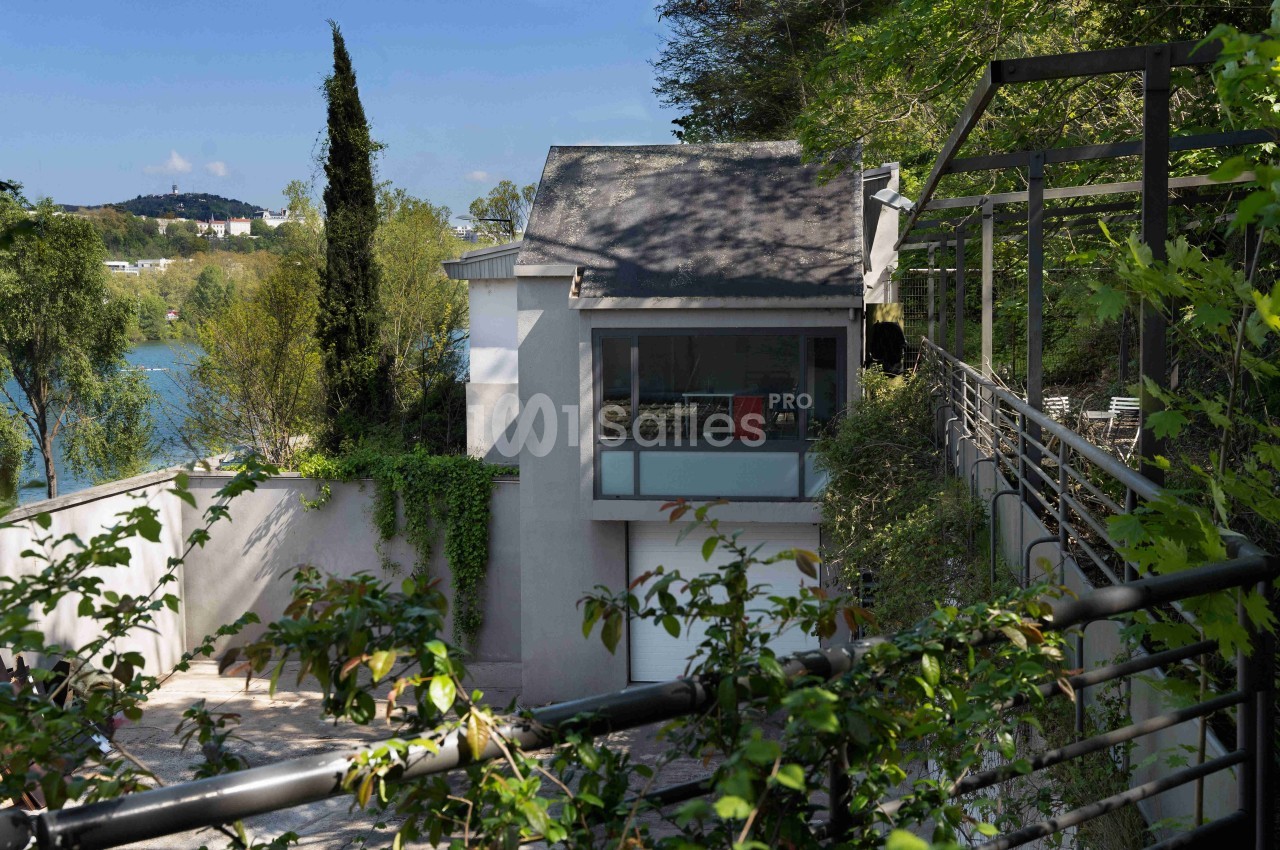 Maison moderne entourée de végétation, avec vue sur un plan d'eau et une terrasse ombragée par une pergola.