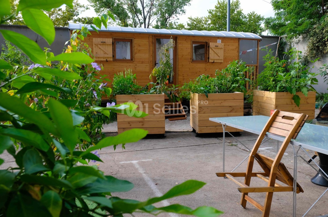Cabane en bois entourée de jardinières avec des plantes, située dans une cour avec une table et une chaise au premier plan.