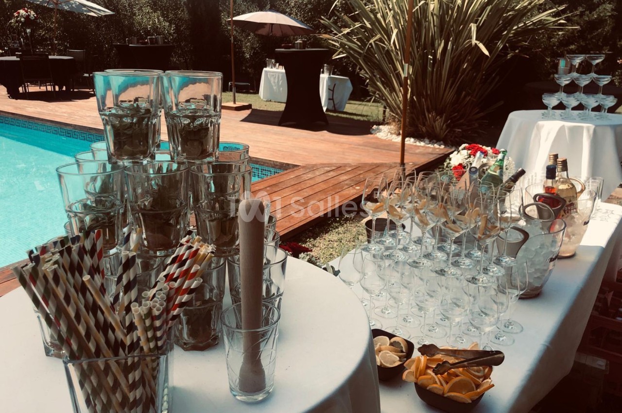 Table dressée près d'une piscine avec verres, pailles, citrons et bouteilles pour un service de boissons en extérieur.
