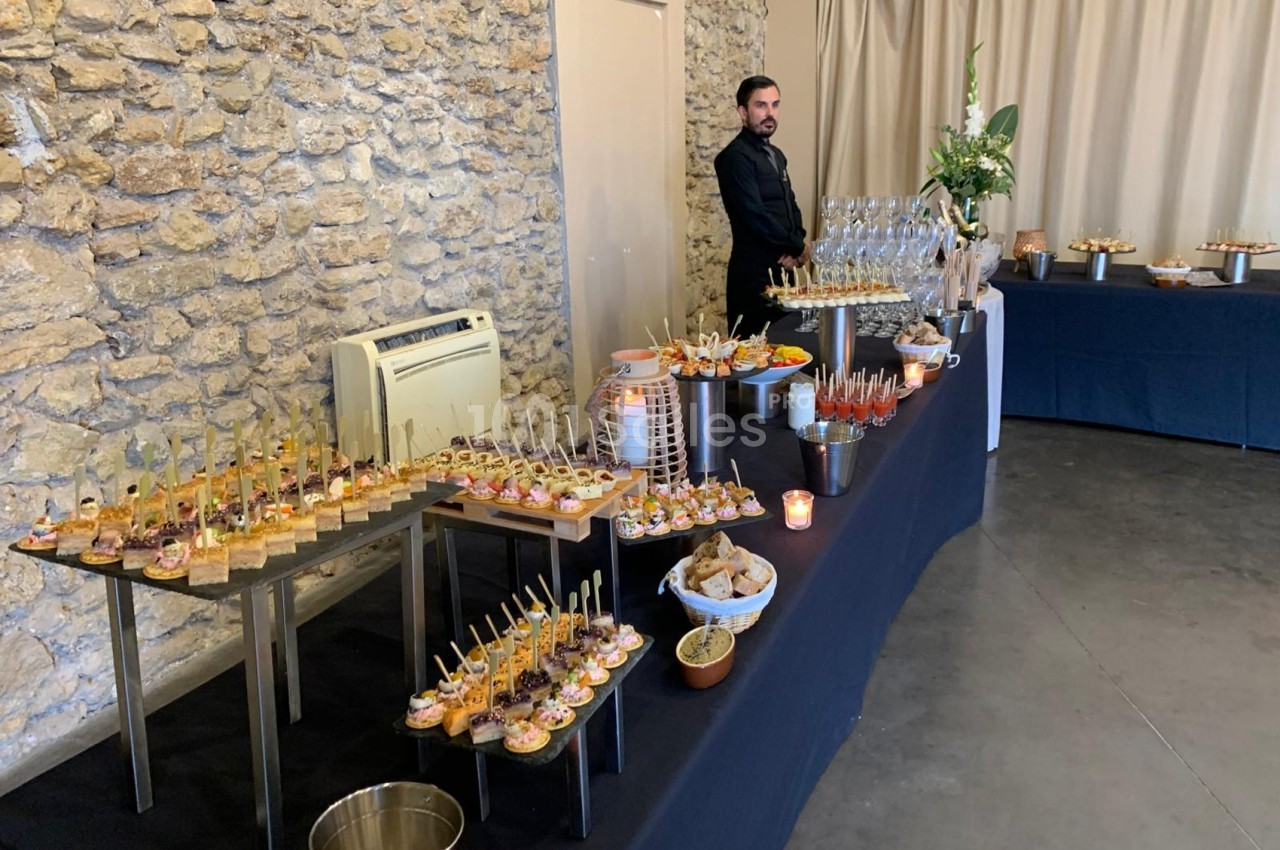 Buffet de réception avec amuse-bouches variés, verres et décor sobre dans une salle aux murs en pierre.