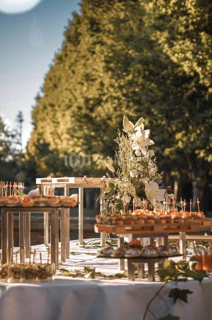 Table garnie de bouchées apéritives et décorée de fleurs blanches, installée en extérieur sous des arbres.