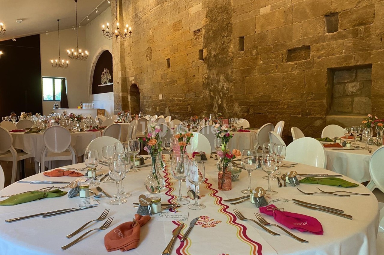Salle de réception décorée pour un événement, avec tables rondes dressées, nappes blanches et arrangements floraux.